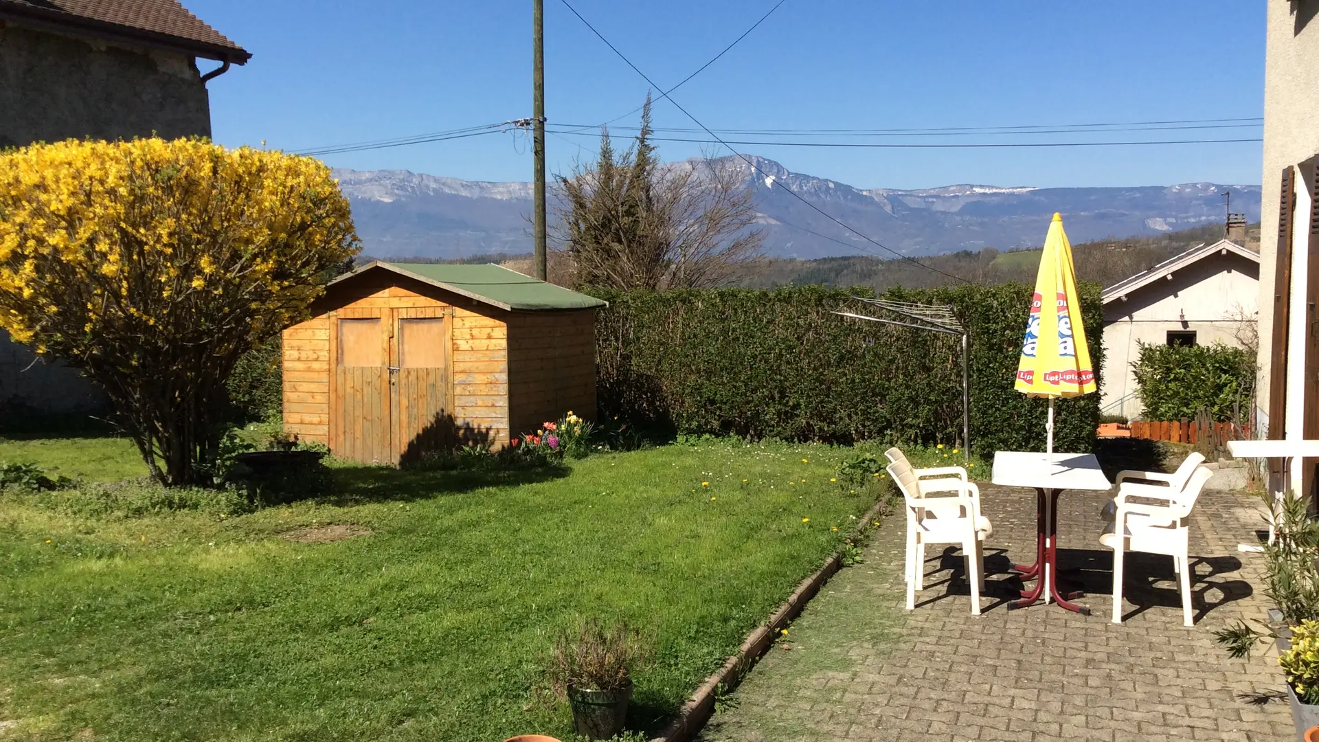 Le jardin est composé d'une pelouse, d'un chemin en gravier et d'un abri de jardin. Une table de jardin avec des chaises est installée pour se détendre.