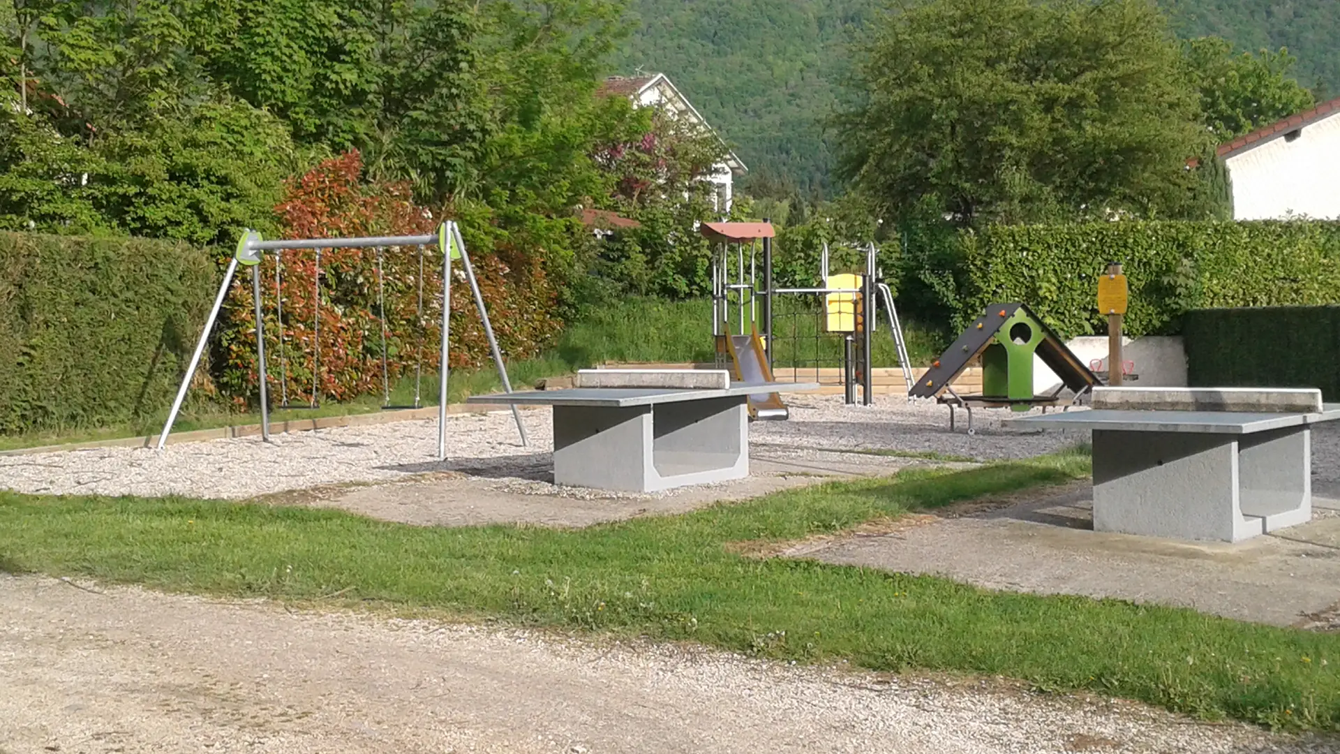 Un espace de jeux en plein air équipé de balançoires suspendues à des chaînes, de modules de jeux en bois pour grimper et ramper et de tables de ping-pong. L'aire de jeux est bordée d'arbres et d'arbustes.