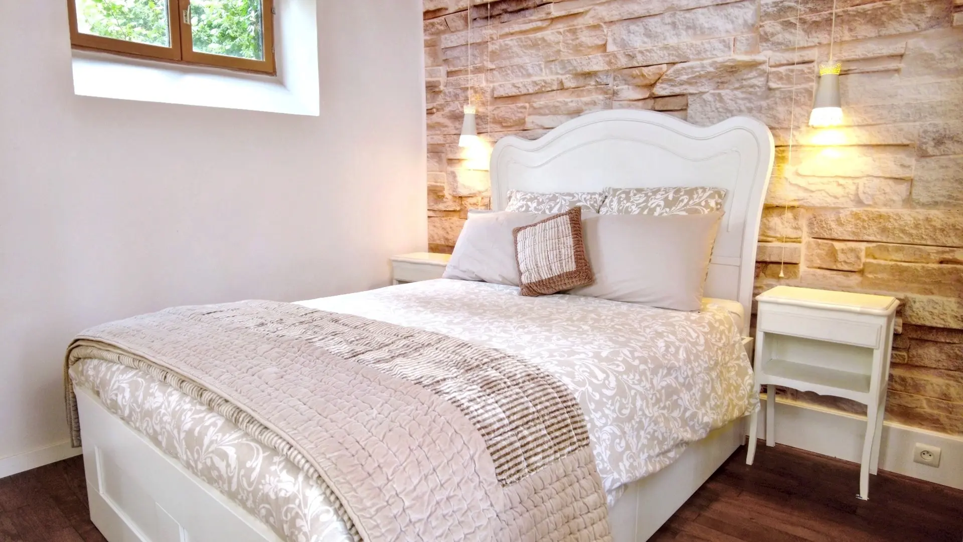 Une chambre à coucher chaleureuse et lumineuse avec un lit double au centre. Le lit est doté d'une tête de lit blanche ornée de moulures et est habillé d'une literie claire, comprenant une couette à motifs beiges et une couverture matelassée de couleur taupe. Au-dessus des oreillers, un coussin décoratif texturé de couleur terre cuite est posé. De chaque côté du lit, il y a une petite table de chevet blanche.