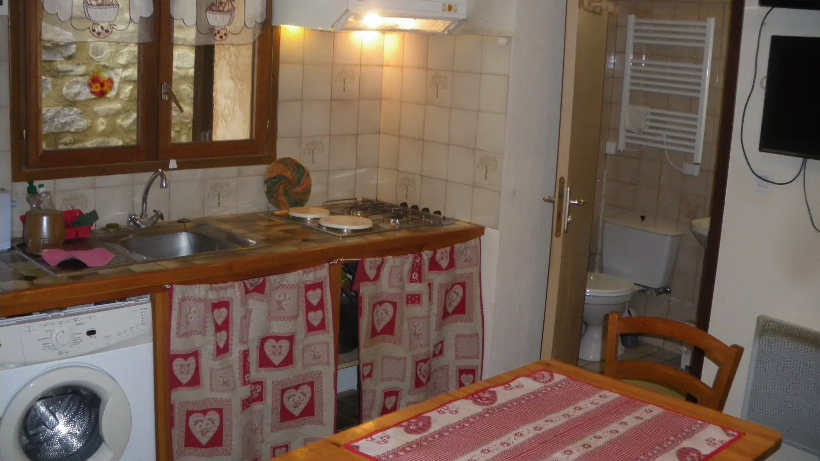 Coin séjour avec cuisine, table à manger, évier, machine à laver, carrelage au mur, ameublement bois, porte vers salle de bains