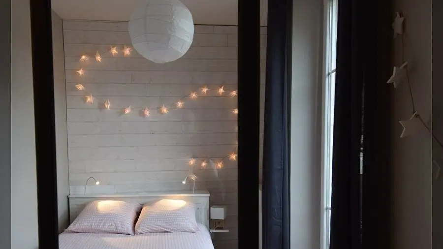 Vue à travers la verrière d'une chambre cosy, avec un lit double et au fond un mur en bois décoré de guirlandes lumineuses en forme d’étoile.