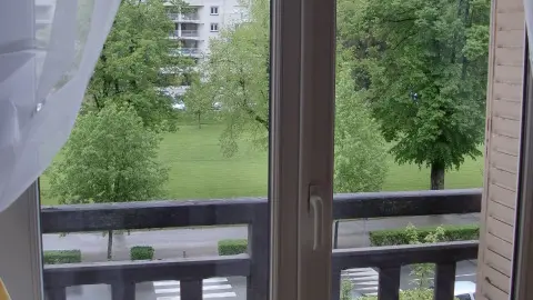 Cette photo montre une vue depuis une fenêtre donnant sur un balcon. À travers des rideaux blancs et voilés, on aperçoit le parc d’Uriage.