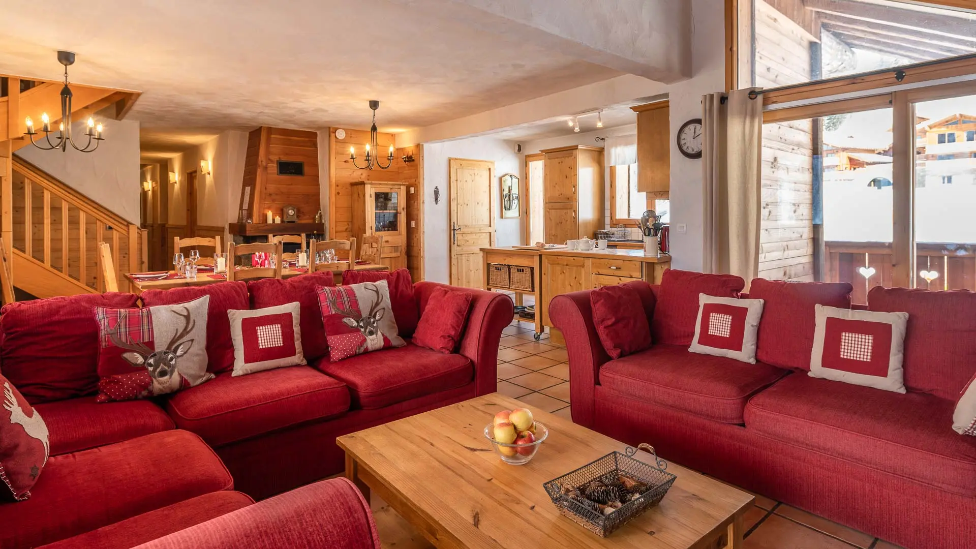 Coin salon avec un grand canapé d'angle rouge et 1 autre canapé, une table basse en bois, puis à l'arrière plan coin cuisine avec une grande table à manger pour 8 personnes situé à côté de la cheminée. Aperçu de l'escalier menant à l'étage où se trouve les chambres