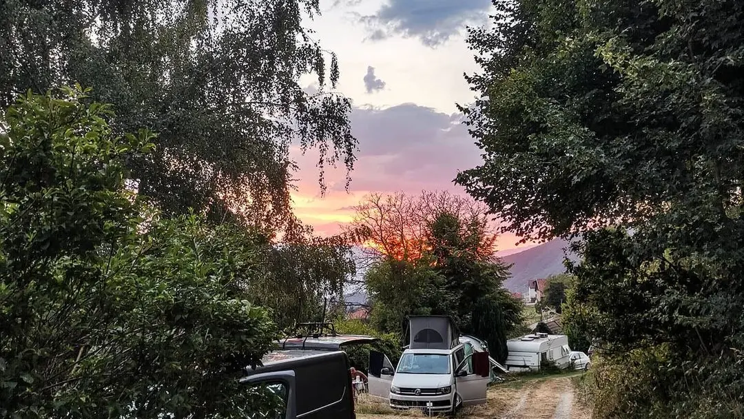 La photo montre un chemin de terre bordé d'arbres, menant à une petite aire de camping où plusieurs vans aménagés sont stationnés. Le ciel est illuminé par les couleurs chaudes d'un coucher de soleil.