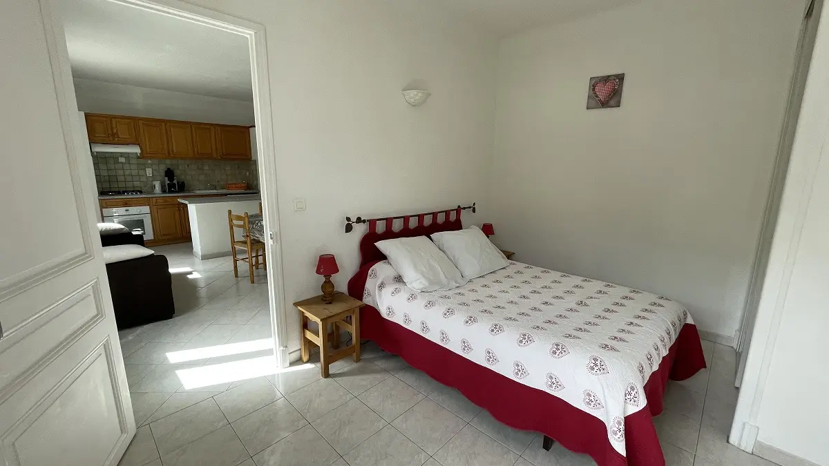 Chambre à coucher lumineuse avec lit double, sol carrelé, ameublement bois, porte vers séjour