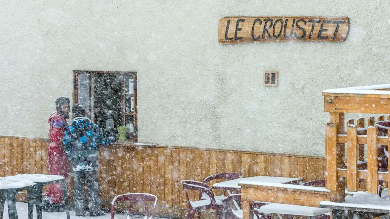 Detail of the restaurant's facade with the wooden sign Le Croustet under the snow, tables and chairs