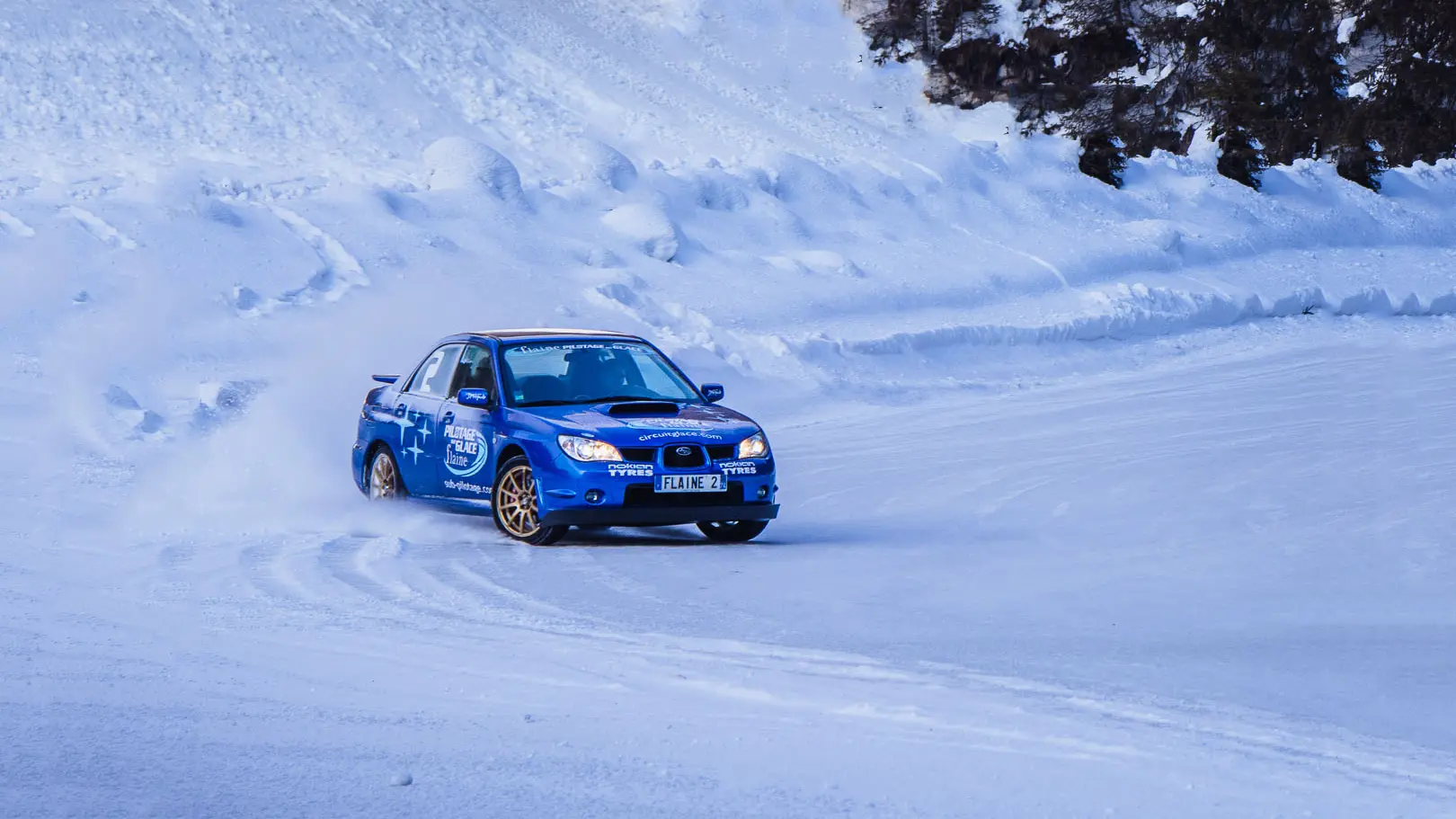 Cette image montre une voiture de rallye bleue en train de déraper sur un circuit de glace à Flaine. La voiture soulève une fine poudre de neige en négociant un virage.
