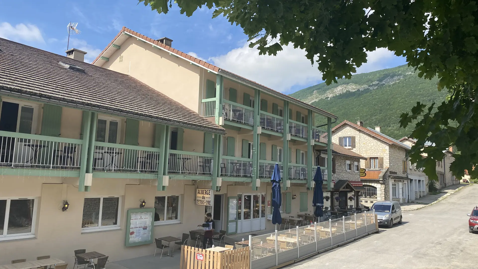 Façade avec hôtel et terrasses aux étages. Restaurant au rez-de-chaussée avec deux terrasses, parasols bleus fermés, tarifs affichés