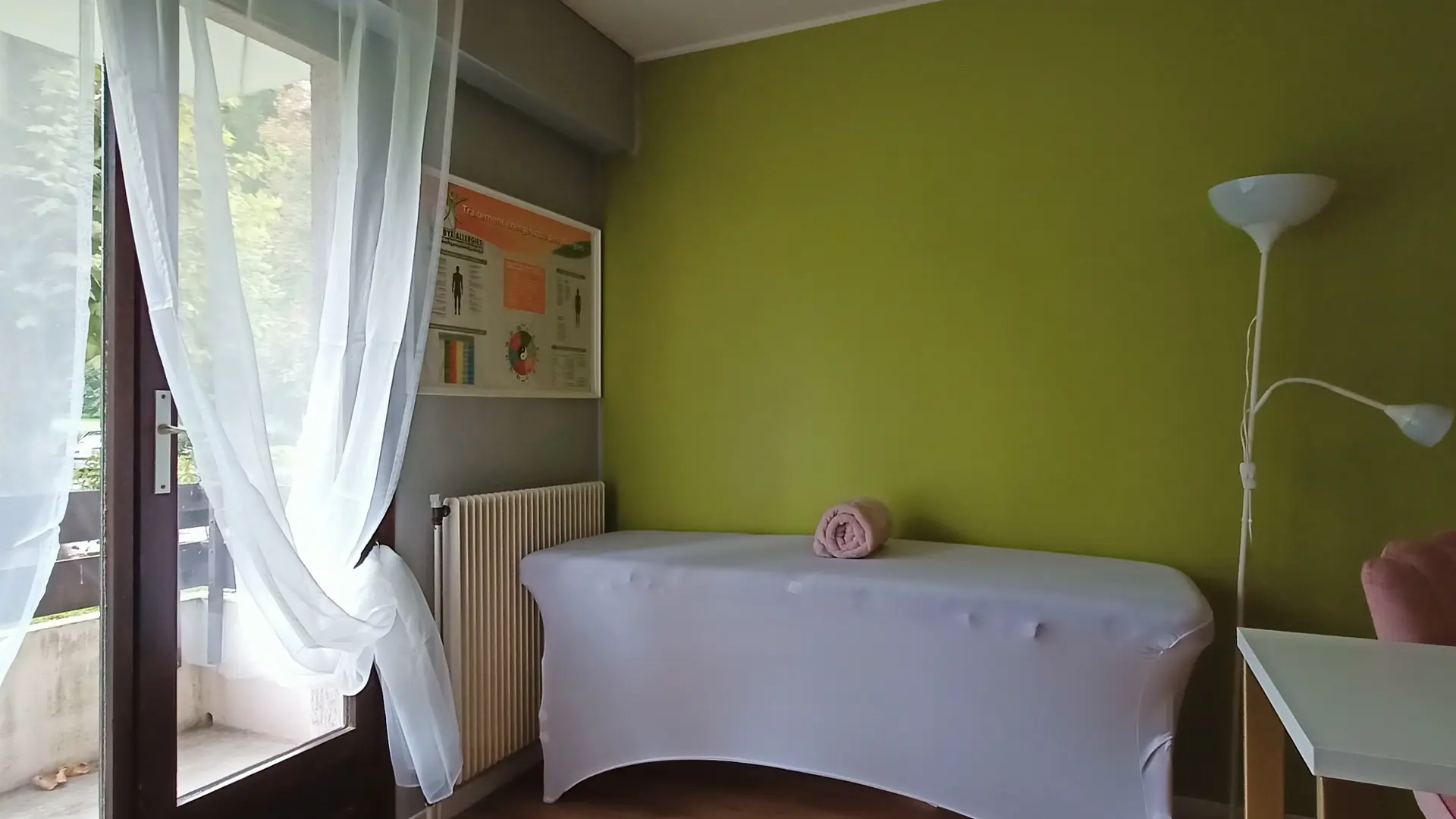 C'est une pièce aux murs vert olive et gris, avec un canapé de massage recouvert d'une housse blanche, une table blanche et une lampe sur pied dans un coin, et une porte-fenêtre avec des rideaux transparents.