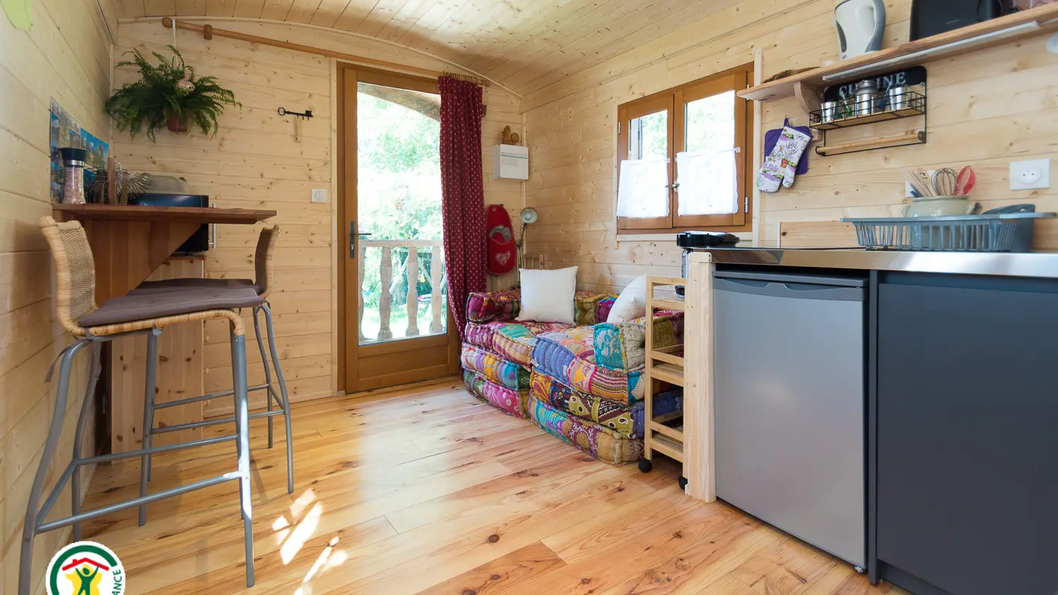 Un petit espace de vie avec des murs en bois clair et un plancher en bois, comprenant une porte-fenêtre menant à l'extérieur, un coin cuisine avec des rangements sombres et un réfrigérateur argenté, et des tabourets de bar dans l'angle opposé.