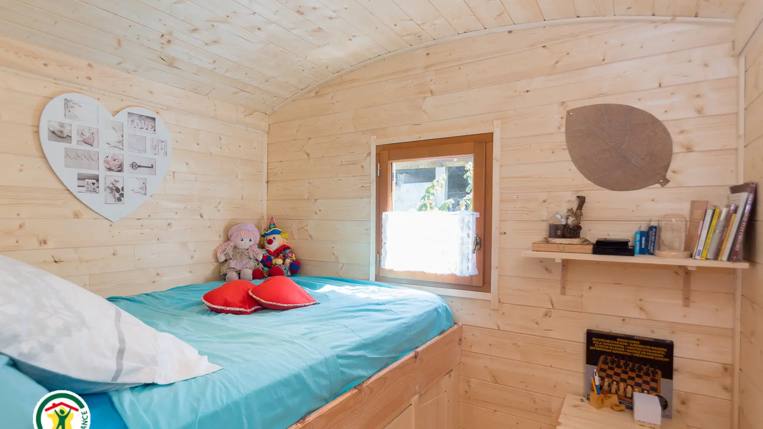 Un espace chambre compact et mansardé avec des murs en bois clair, contenant un lit recouvert d'une couette turquoise avec un oreiller blanc, une petite fenêtre à cadre marron et une étagère fixée au mur.