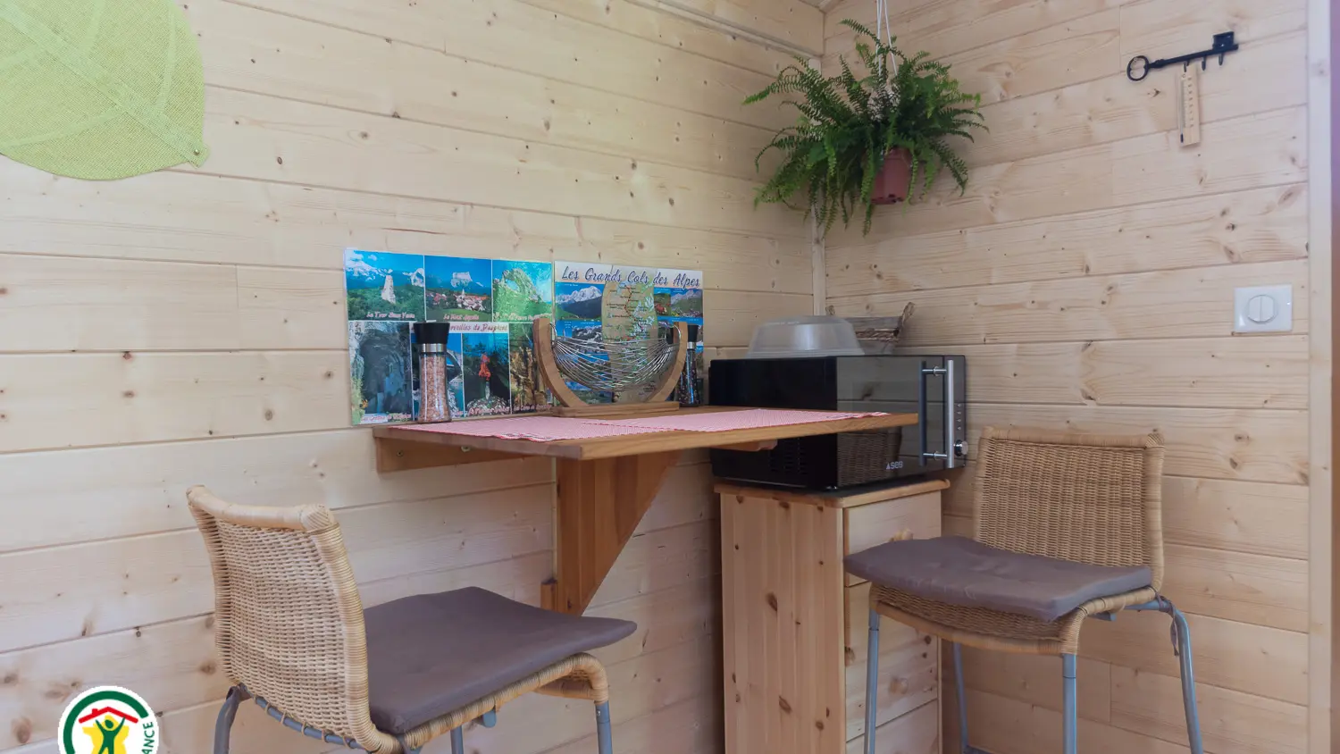 Un coin repas dans un petit espace aux murs et plancher en bois clair, composé d'une petite table pliante marron fixée au mur et de deux chaises en face du coin cuisine.