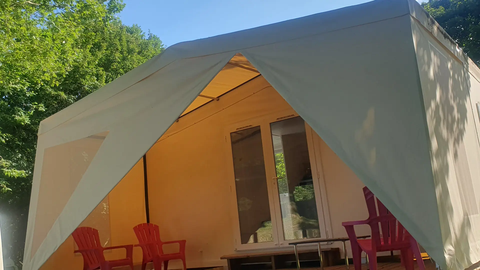 Un hébergement insolite de type lodge, caractérisé par une structure légère avec une toiture tendue en tissu clair. Une terrasse en bois aménagée avec deux fauteuils donne accès à l'intérieur du logement par deux larges portes vitrées.