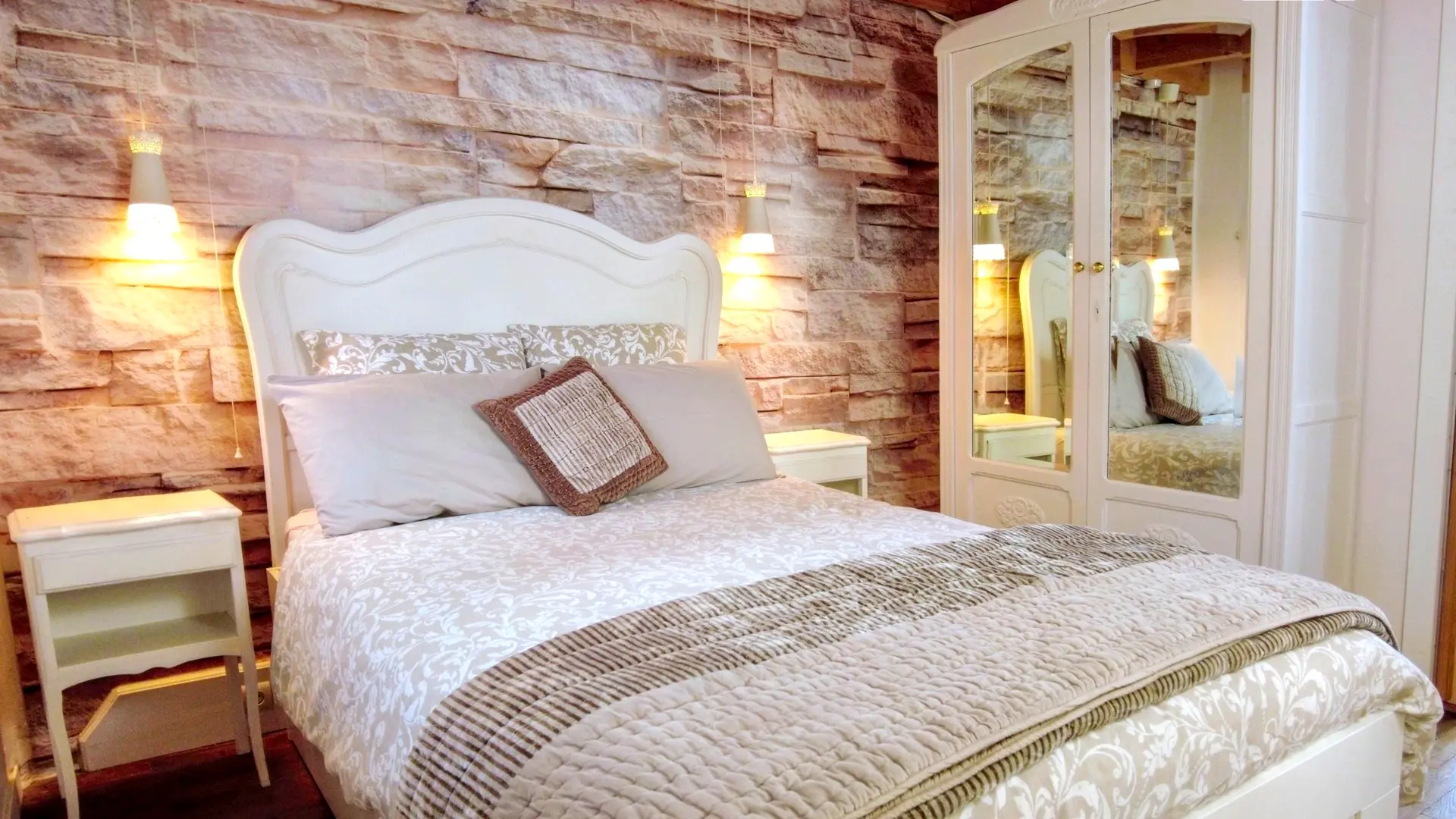 Une chambre à coucher chaleureuse et lumineuse avec un lit double au centre. Le lit est doté d'une tête de lit blanche ornée de moulures et est habillé d'une literie claire, comprenant une couette à motifs beiges et une couverture matelassée de couleur taupe. Au-dessus des oreillers, un coussin décoratif texturé de couleur terre cuite est posé. De chaque côté du lit, il y a une petite table de chevet blanche. À droite, une grande armoire blanche à deux portes est visible. Les portes sont équipées de miroirs rectangulaires qui reflètent la pièce et la lumière.