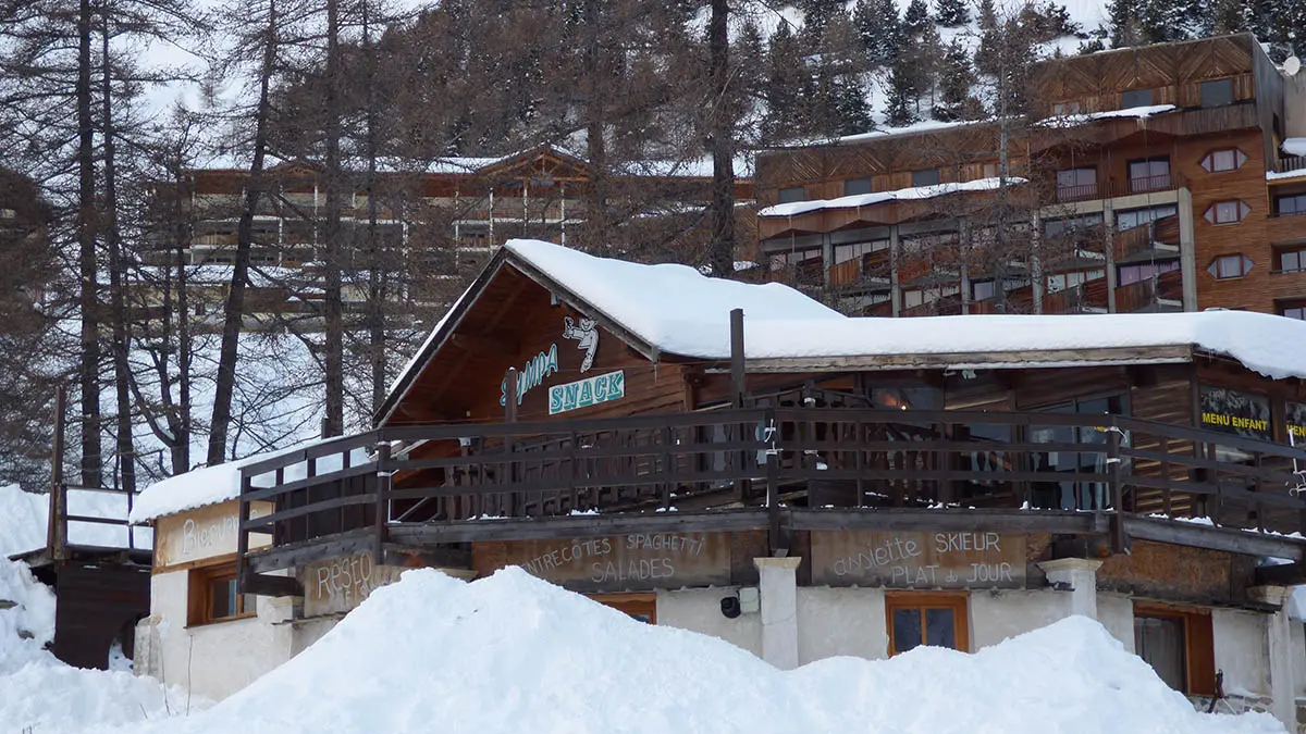 Vue d'ensemble hivernale du restaurant en bois, situé devant des immeubles de la station de ski, en haut d'un bâtiment sur deux niveaux, terrasse. Panneau Sympa Snack sur la devanture. Montagnes enneigées en arrière-plan.