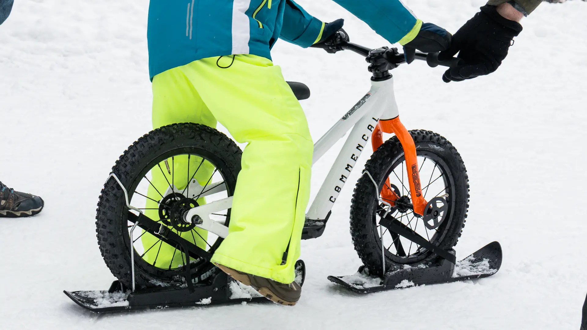 Un enfant portant un pantalon jaune fluo et une veste bleue est assis sur un vélo-ski blanc. On voit bien les patins noirs fixés sous les roues pour glisser sur la neige.