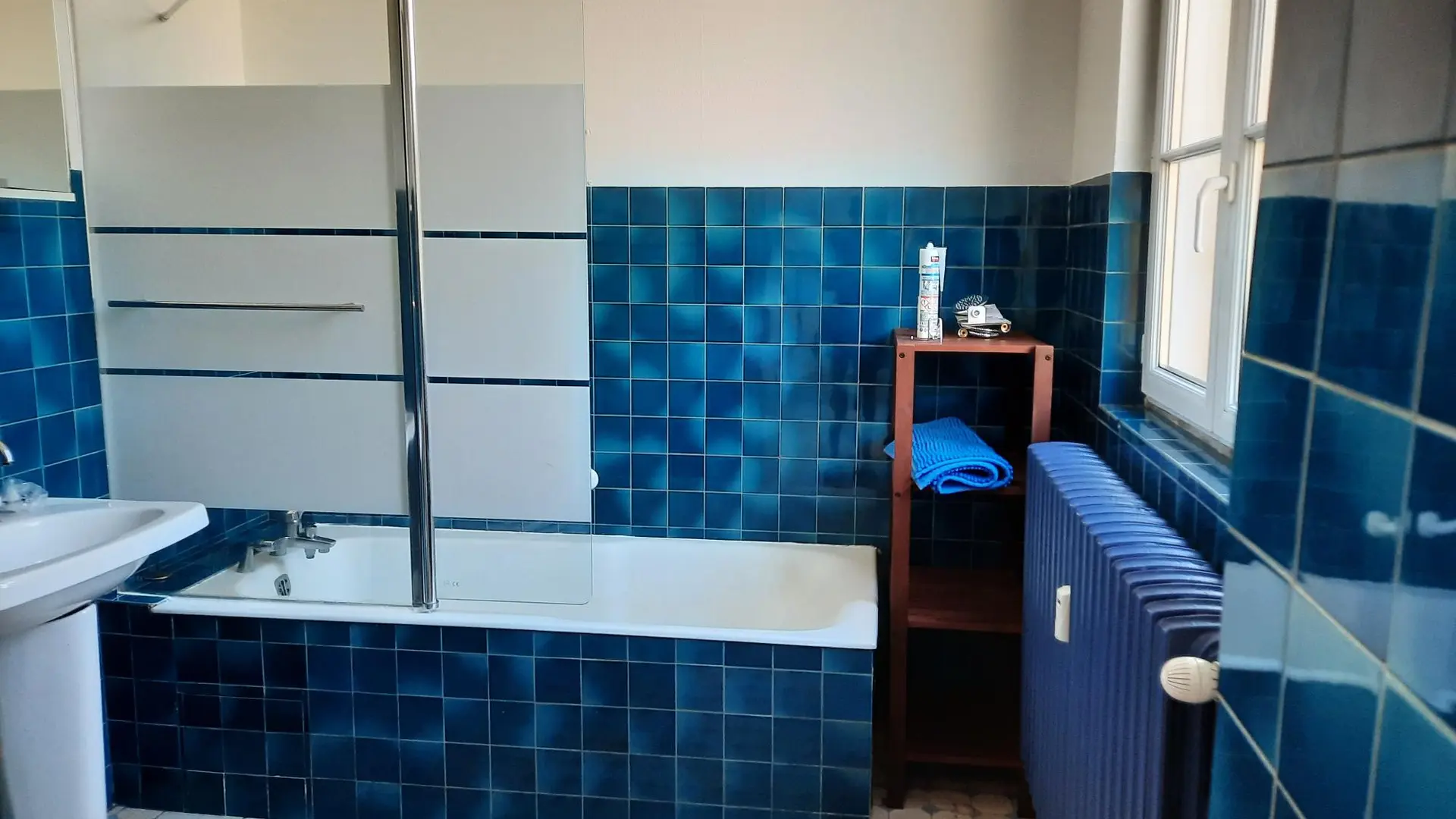 Il s'agit d'une salle de bain carrelée de carreaux bleus et blancs, équipée d'une baignoire avec un pare-douche, un lavabo sur pied, un meuble étagère en bois et un radiateur bleu sous une fenêtre.