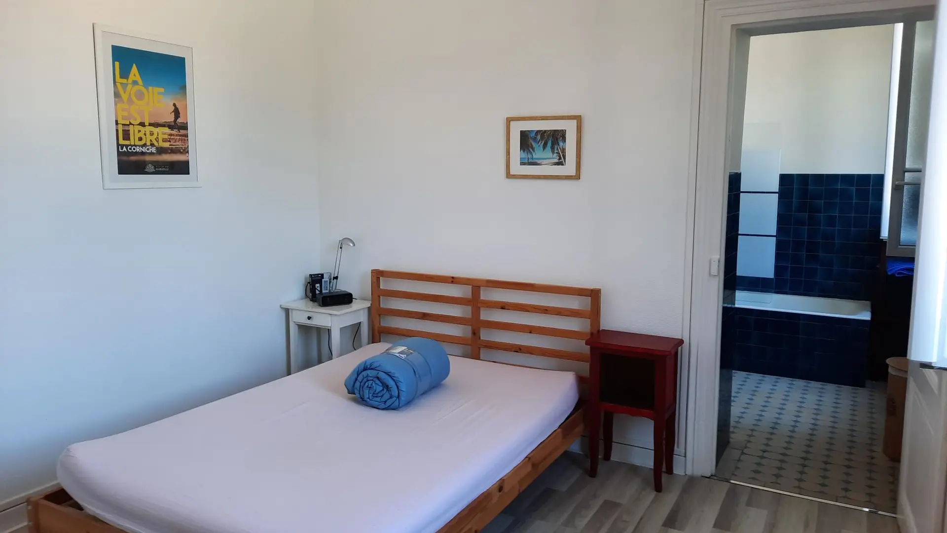 Une chambre avec un lit double sur une armature en bois, une table de chevet et des décorations murales, et une salle de bains visible par une porte ouverte au fond.