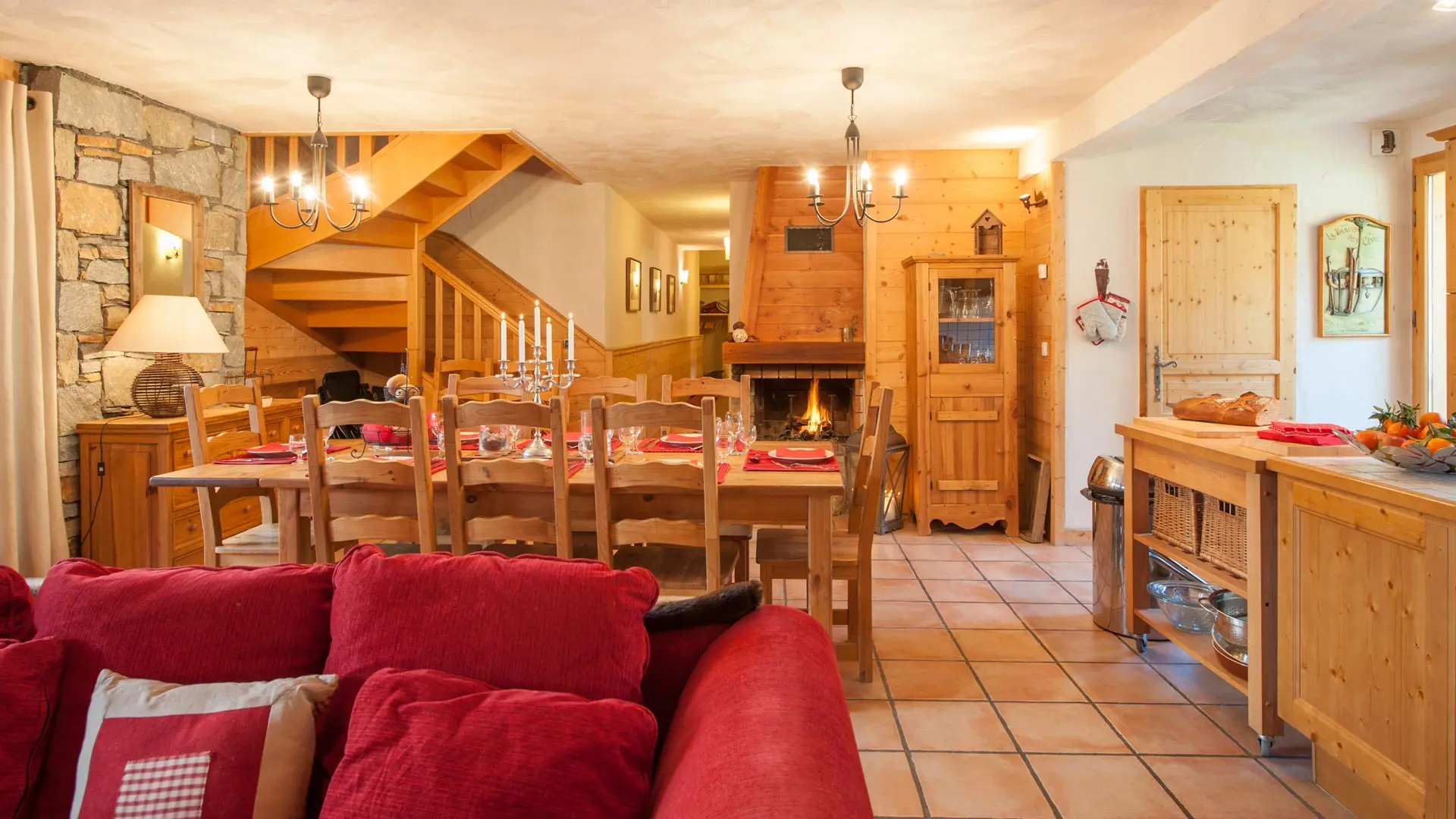 Grande pièce de vie, grande table à manger en bois pour 8 personnes, cheminée, commode avec tiroirs et luminaire, grand escalier en bois, meuble de rangement vaisselle. Salon avec un grand canapé rouge.