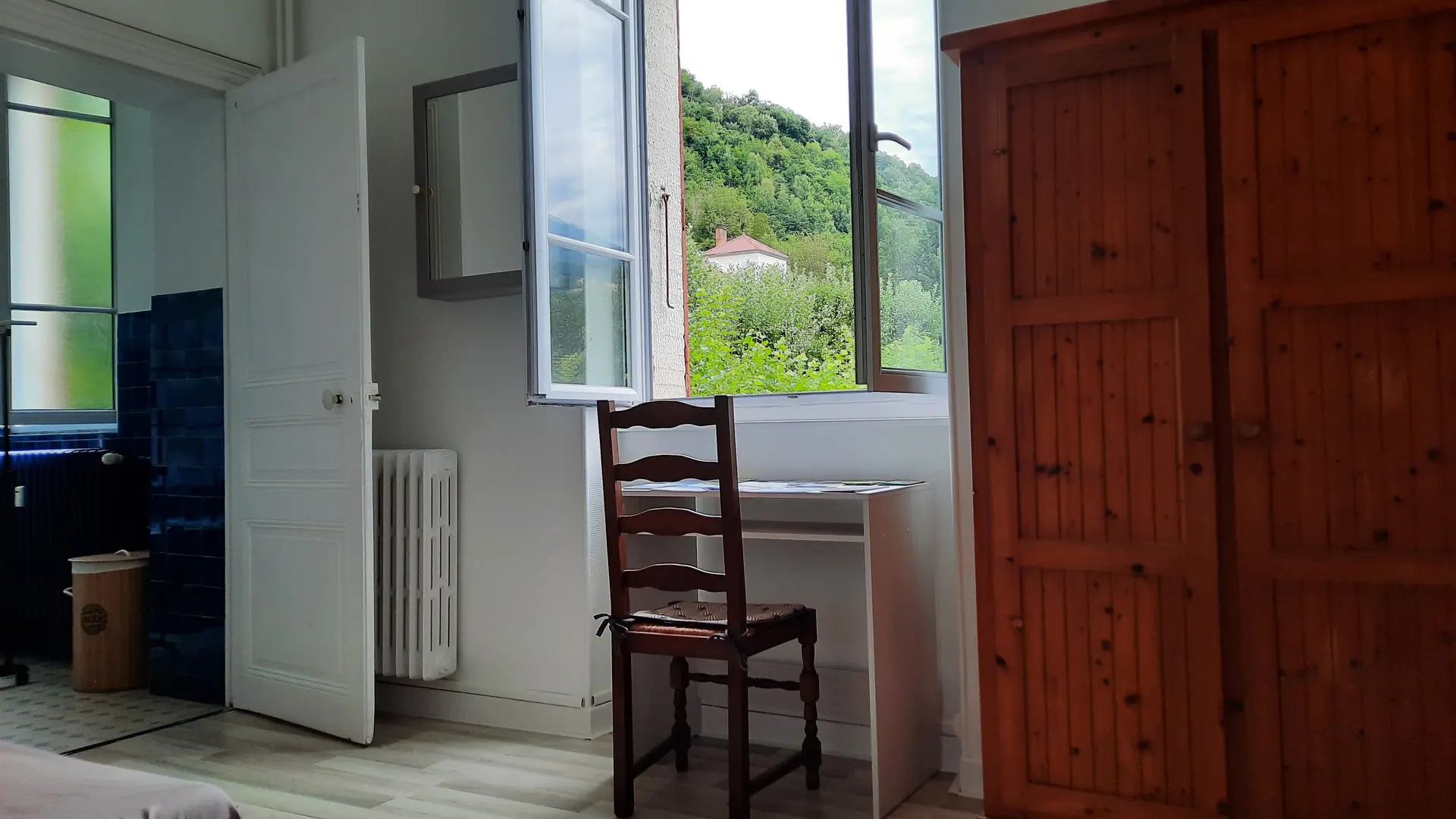 C'est une chambre meublée d'une grande armoire en bois, d'un bureau avec une chaise devant une fenêtre avec une vue sur de la végétation à l'extérieur.