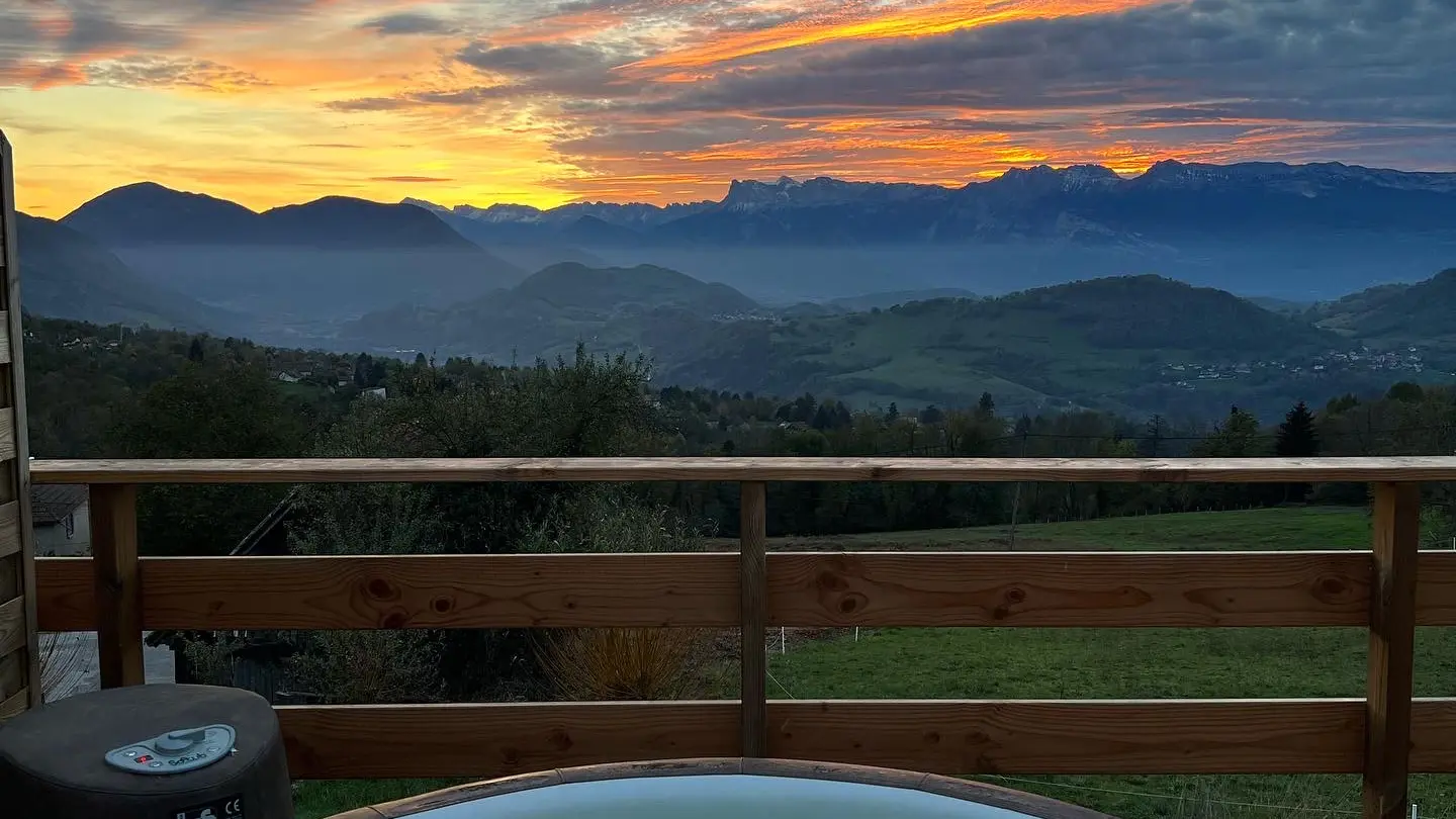 La photo montre un spa rond situé sur une terrasse en bois, offrant une vue panoramique sur une chaîne de montagnes à l'horizon. Le ciel est illuminé par les couleurs vives d'un coucher de soleil.