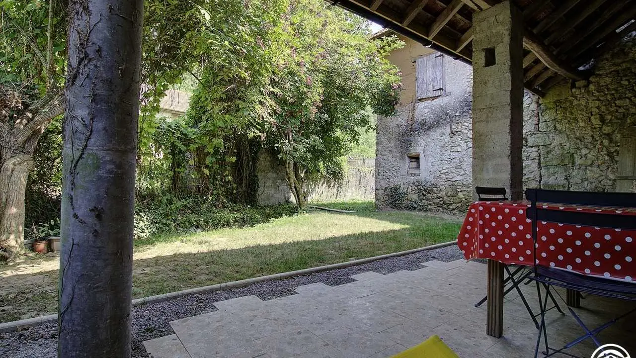 Vue de la terrasse couverte, son petit jardin ombragé, bordé par la petite rivière