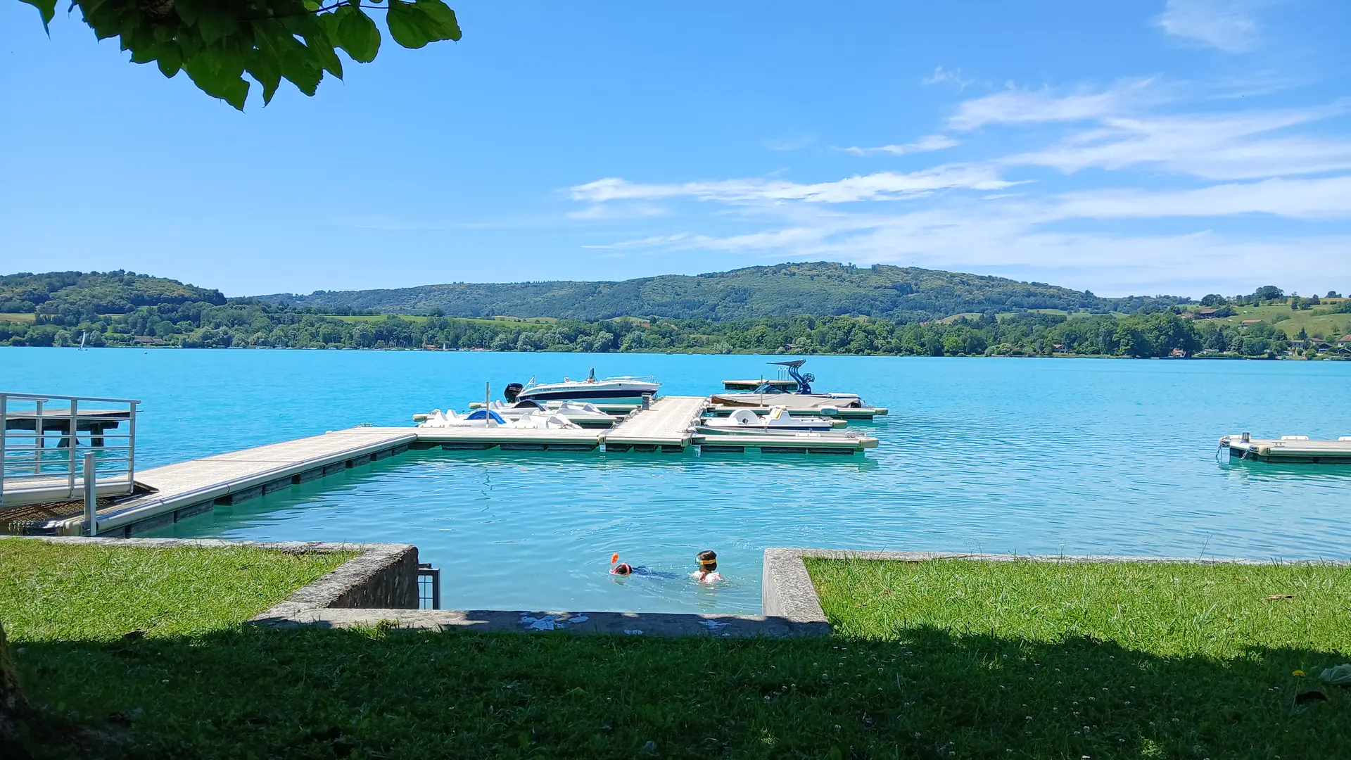 Au premier plan, un liseré d'herbe ouvre sur les eaux turquoises d'un lac scintillant sous un soleil radieux. Un ponton où sont amarrés bateaux et pédalos invite à l'aventure.