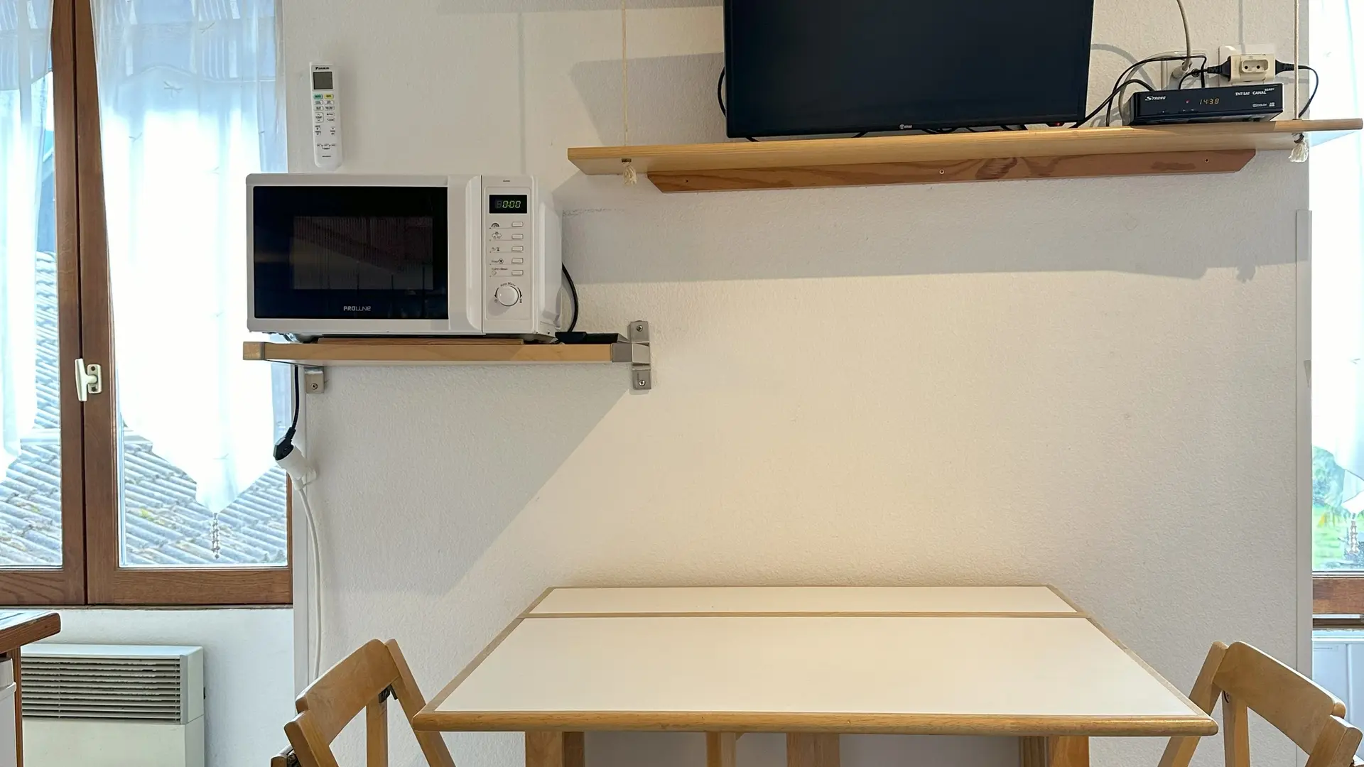 Un espace repas est aménagé avec une table escamotable carrée en bois clair et deux chaises pliantes assorties. Un micro-ondes est fixé au mur, juste au-dessus d'une étagère.