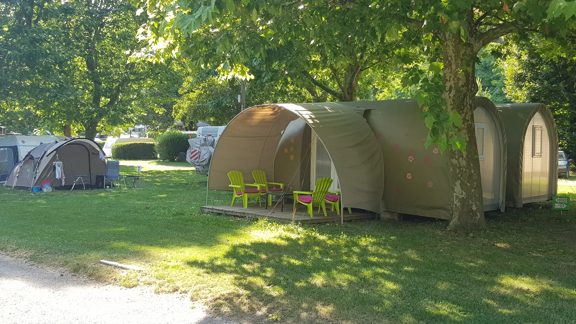 Un logement de plein air de forme atypique avec une toiture tendue et une terrasse en bois, doté de deux grandes baies vitrées et meublé de deux fauteuils.