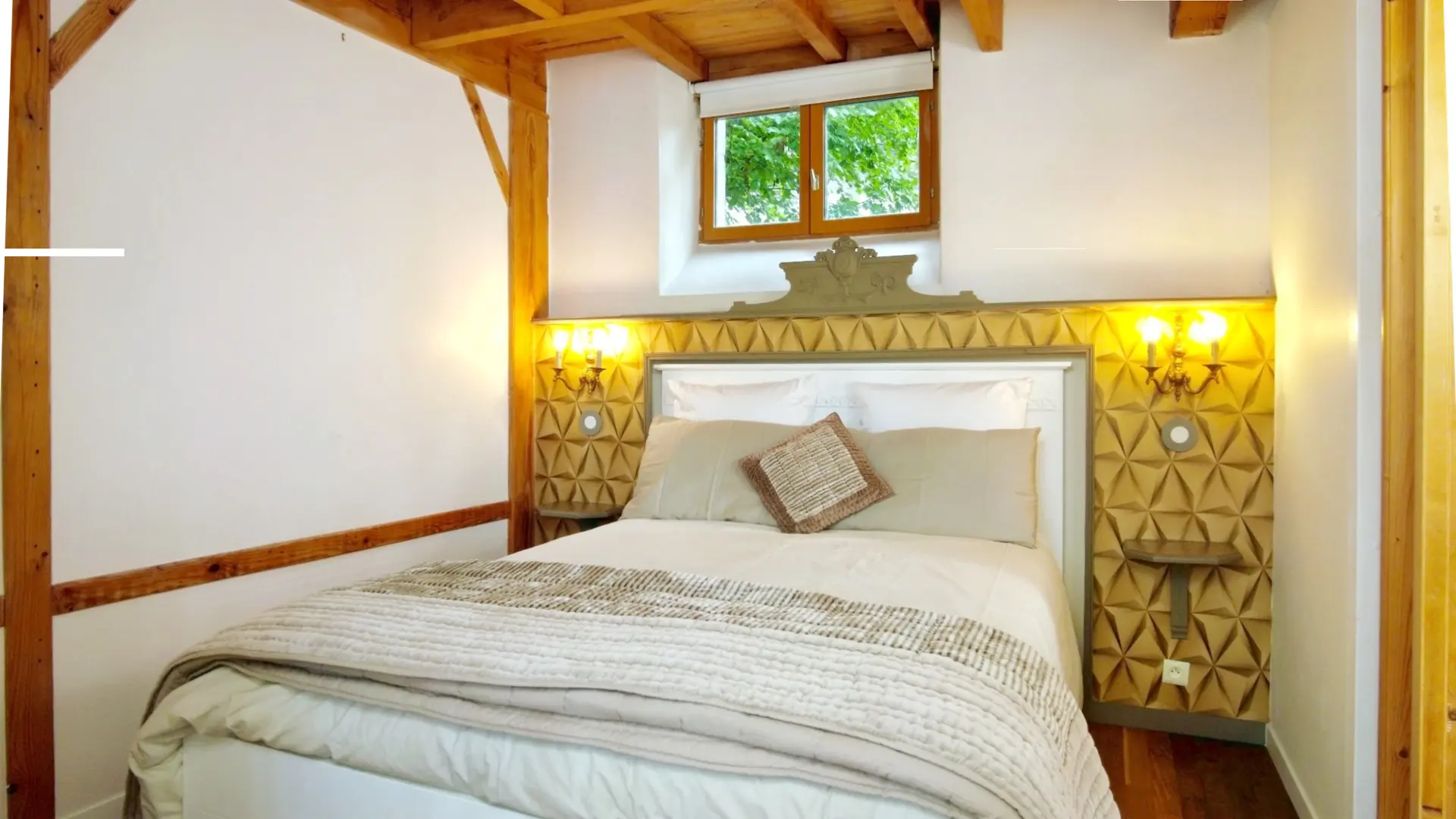 Une chambre à coucher rustique et élégante avec un lit double en son centre. Le lit est doté d'une tête de lit blanche avec un encadrement gris-vert orné de motifs sculptés. Il est habillé d'une couette blanche, d'un plaid matelassé beige et de coussins moelleux, avec un petit coussin décoratif tressé au centre. Le mur derrière le lit est décoré de panneaux géométriques texturés de couleur or clair, et de chaque côté du lit, des appliques murales jaunes diffusent une lumière chaude. Des petites tables de chevet murales sont intégrées aux panneaux. La chambre présente des murs blancs et des poutres en bois apparentes au plafond. Une fenêtre en bois au-dessus de la tête de lit offre une vue sur la verdure extérieure.
