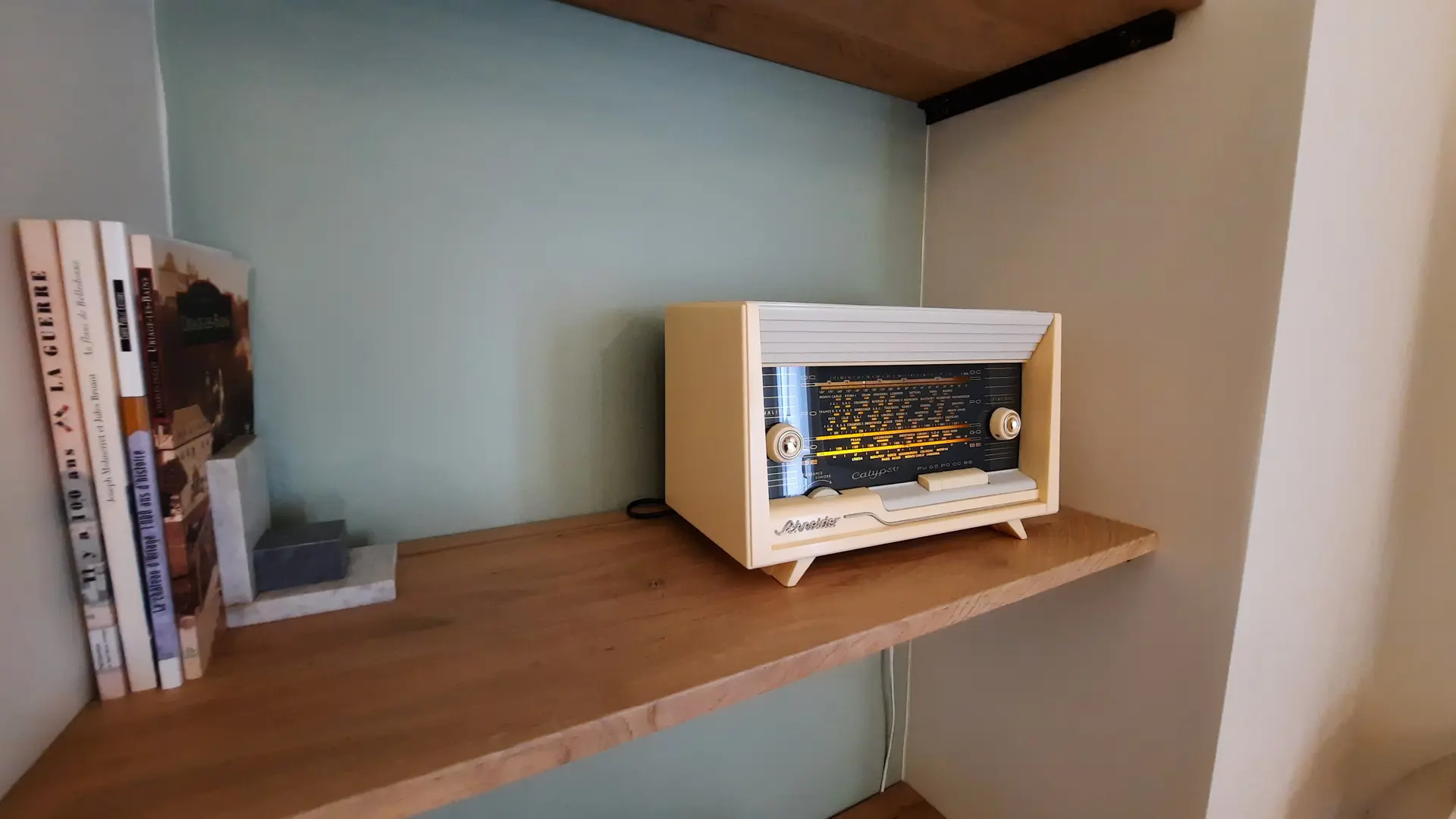 Sur une étagère en bois clair, vous trouverez une radio vintage en bois et des livres reliés, appuyés contre des objets lourds.