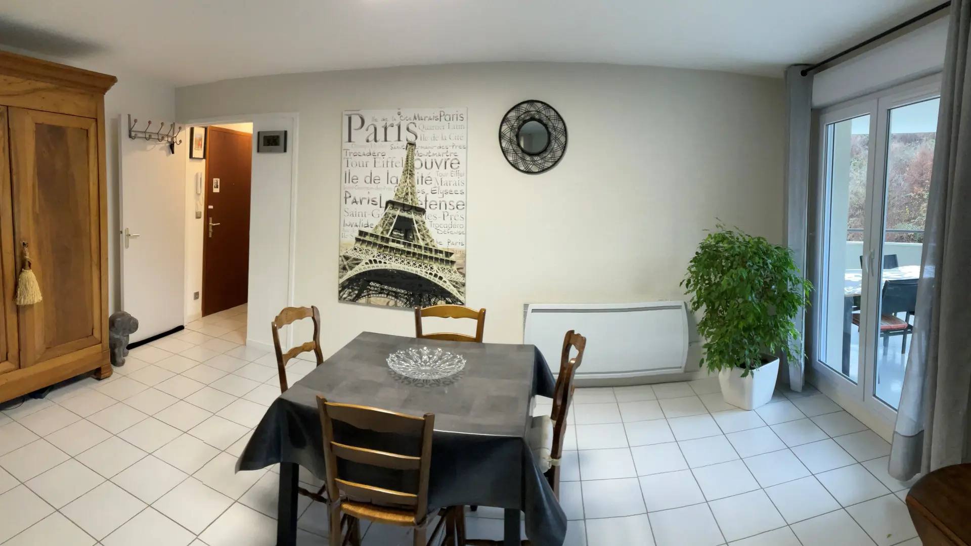 Au centre, une table de salle à manger en bois, recouverte d'une nappe, peut accueillir 4 personnes. Autour de la table, vous trouverez des chaises en bois. Sur un des murs, vous trouverez une armoire et une plante verte. Une porte-fenêtre laisse entrer la lumière naturelle.