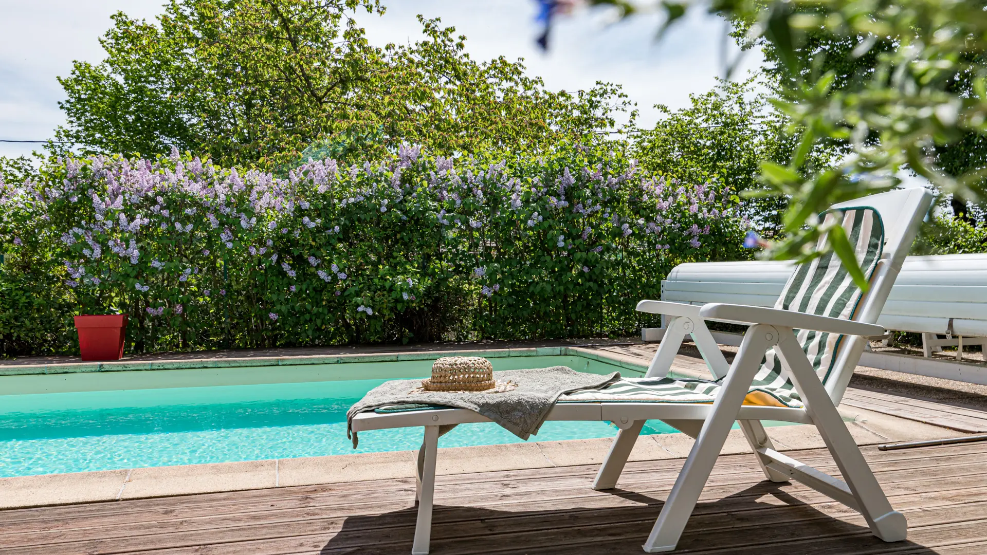 Piscine au sel avec bain de soleil, chapeau et lilas en fleurs dans le jardin arboré.