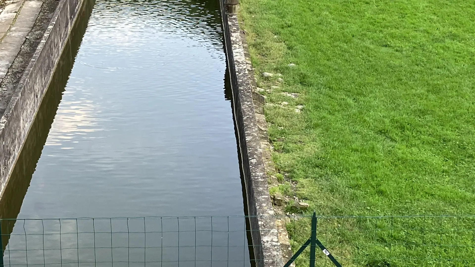 Un étroit canal d'eau traverse ce terrain. Les berges du canal sont recouvertes d'une végétation luxuriante et une petite clôture métallique sépare le cours d'eau d'une pelouse verdoyante.