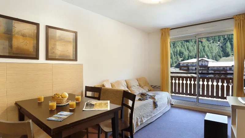 Bright apartment living room with sofa, TV unit, TV, dining table, French doors opening onto a balcony, view of the resort