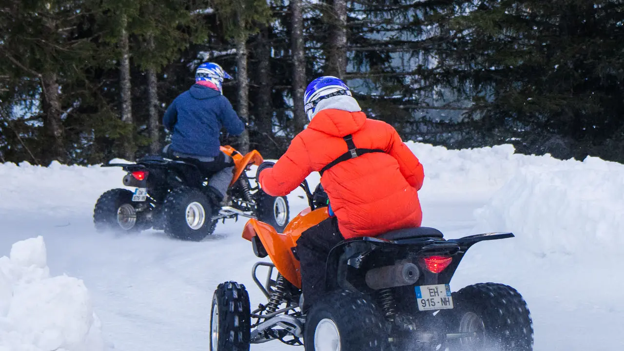 Deux personnes portant des casques et des vêtements d'hiver colorés pilotent des quads orange sur un sentier enneigé bordé de sapins