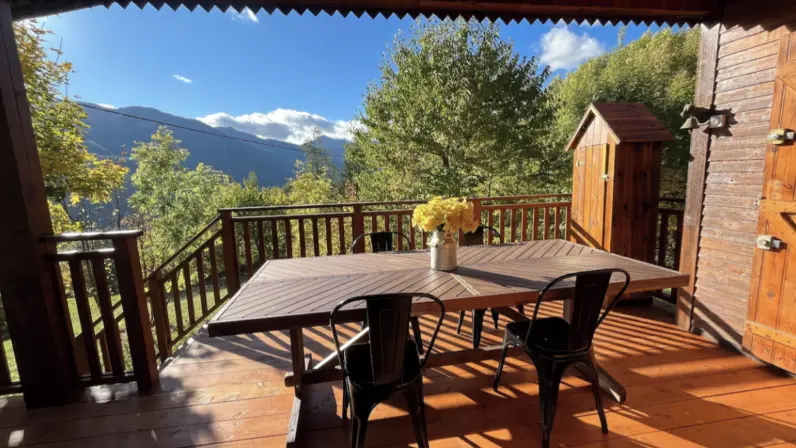 Wooden terrace with a wooden table and iron chairs, small wooden garden shed. Mountain view.