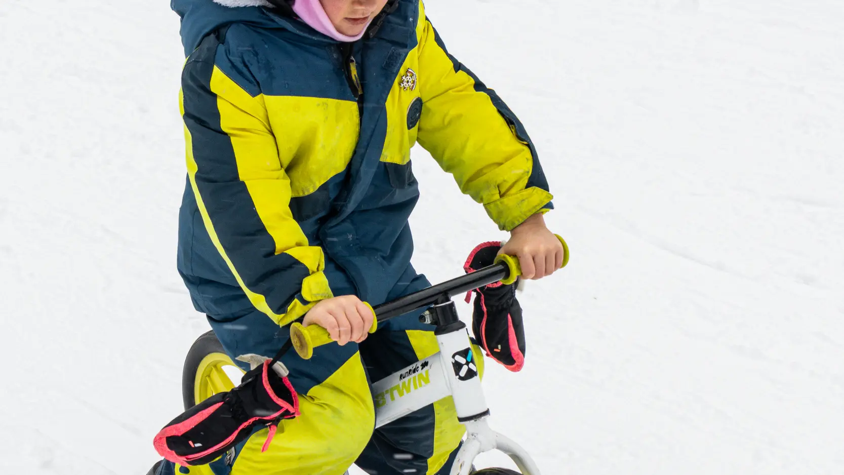 Un jeune enfant portant une combinaison de ski colorée et un bonnet rose en forme de tête d'animal descend une pente de neige sur un petit vélo-ski blanc.
