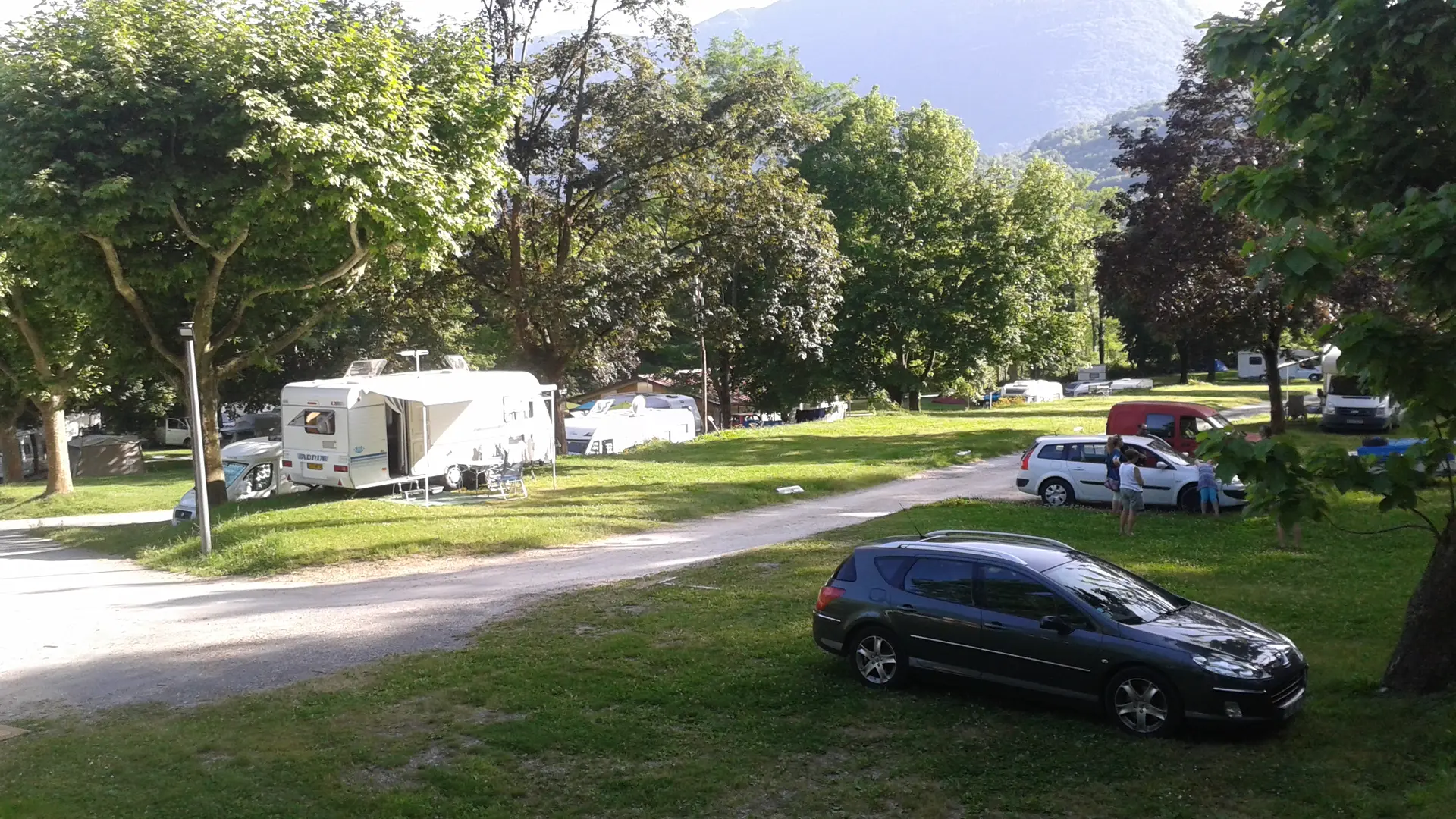 Un espace de camping en plein air comprenant des parcelles individuelles séparées par de la végétation. On y trouve des caravanes, des camping-cars et des voitures stationnées sur un terrain herbeux. L'ensemble est entouré d'arbres et offre une vue sur des collines en arrière-plan.