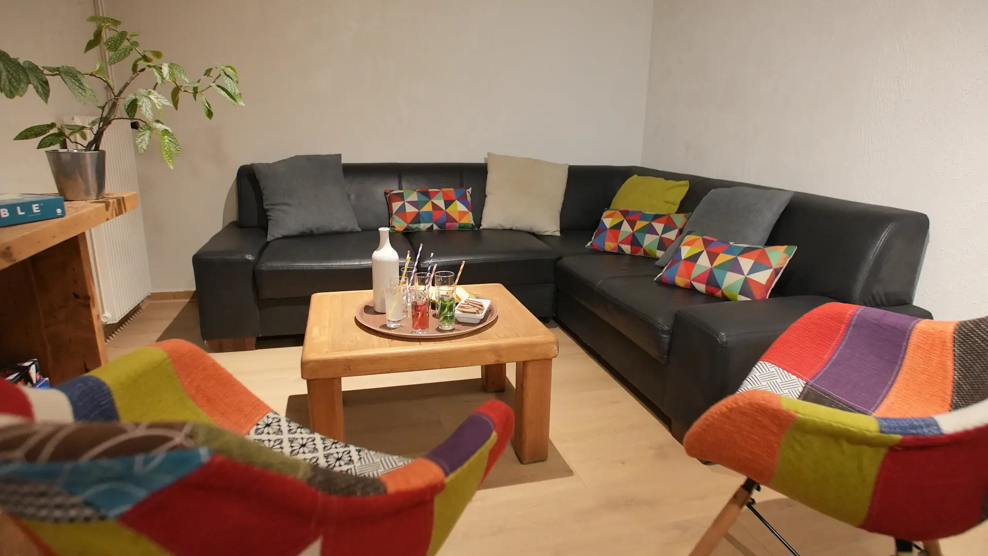 Canapé d’angle avec coussins colorés, deux chaises patchwork et au centre une table avec plateau et verres. Plante décorative