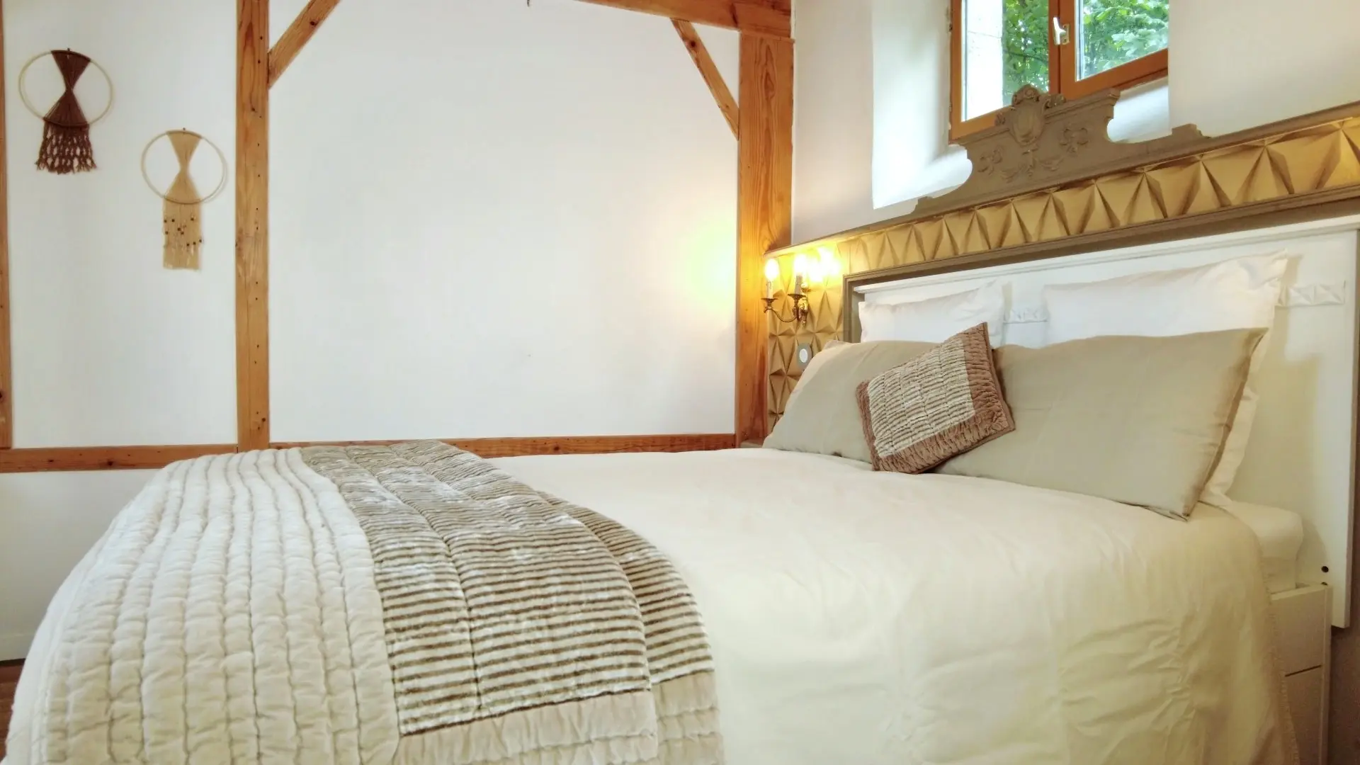 Une chambre à coucher rustique et élégante avec un lit double en son centre. Le lit est doté d'une tête de lit blanche avec un encadrement gris-vert orné de motifs sculptés. Il est habillé d'une couette blanche, d'un plaid matelassé beige et de coussins moelleux, avec un petit coussin décoratif tressé au centre. Le mur derrière le lit est décoré de panneaux géométriques texturés de couleur or clair, et de chaque côté du lit, des appliques murales jaunes diffusent une lumière chaude. Des petites tables de chevet murales sont intégrées aux panneaux. La chambre présente des murs blancs et des poutres en bois apparentes au plafond. Une fenêtre en bois au-dessus de la tête de lit offre une vue sur la verdure extérieure.