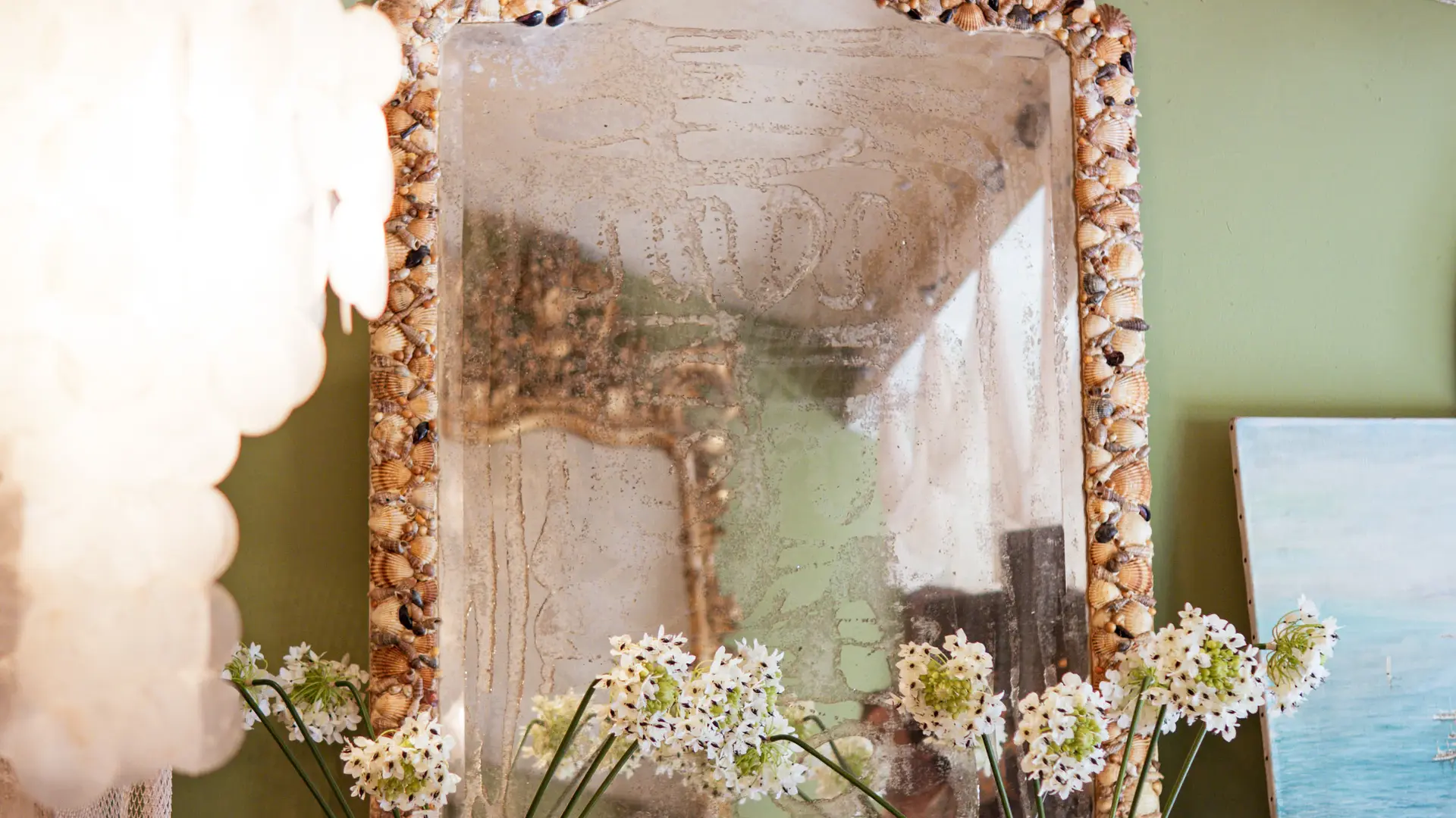 Un miroir encadré de coquillages se reflète dans un décor poétique mêlant fleurs, peintures et meubles anciens