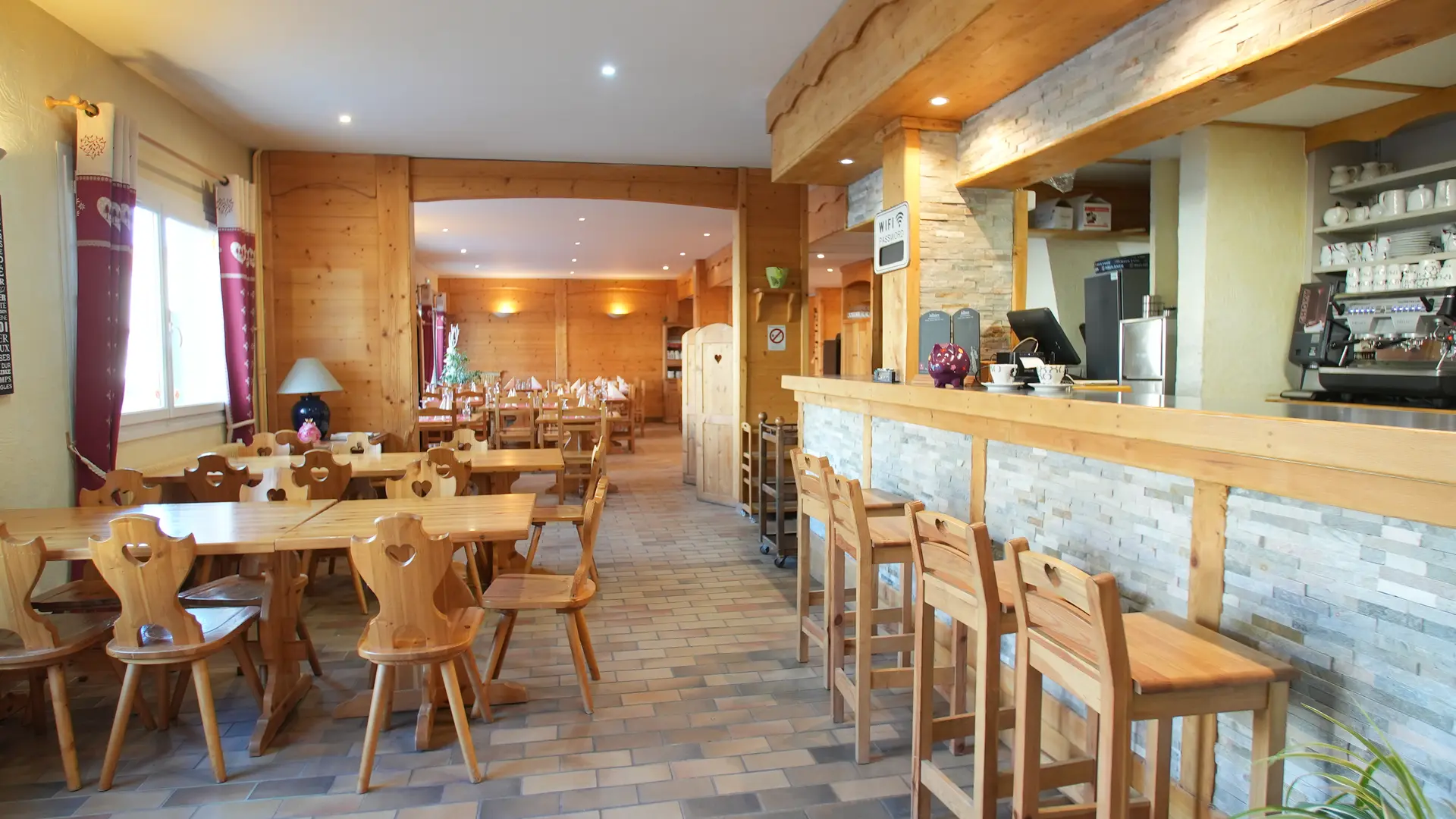 Quatre tabourets sont alignés devant le bar à droite tandis que deux tables avec chaises sont disposées à gauche, le tout en bois. Une fenêtre apporte la lumière naturelle, une cloison ouverte sépare la salle de restauration où les tables sont dressées