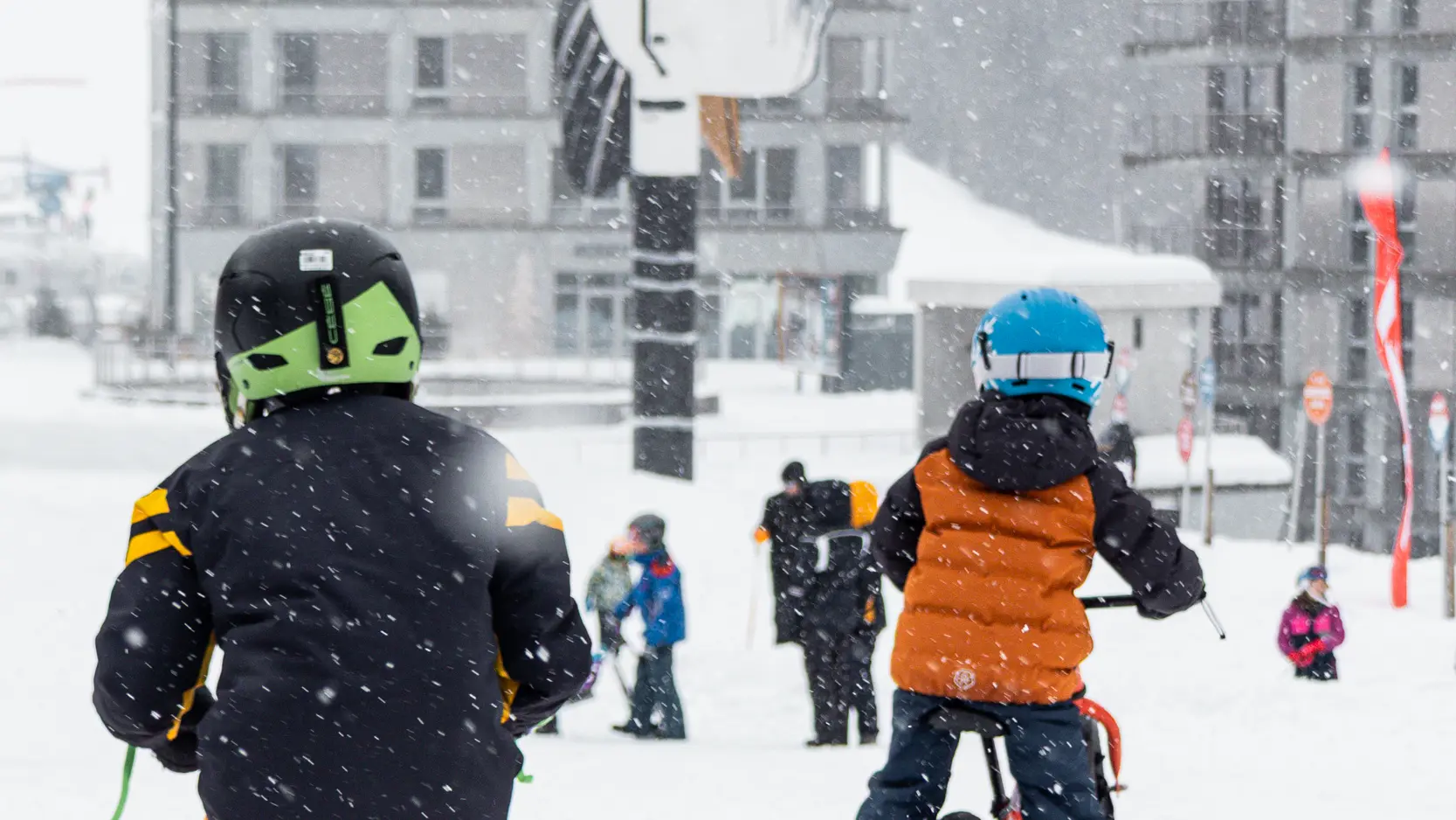 Deux enfants de dos, équipés de casques et de vêtements de ski colorés, font du vélo-ski sur une piste enneigée.