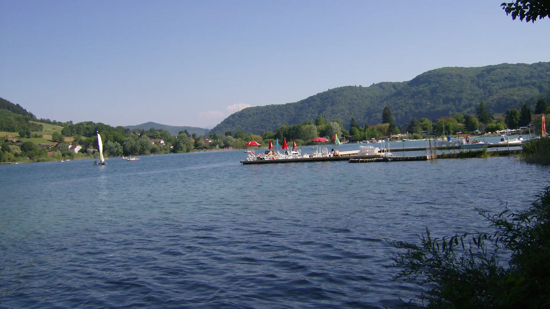 Une vue sur le lac, depuis les rives du lac de Paladru où l'on voit des transats sur un ponton au loin.