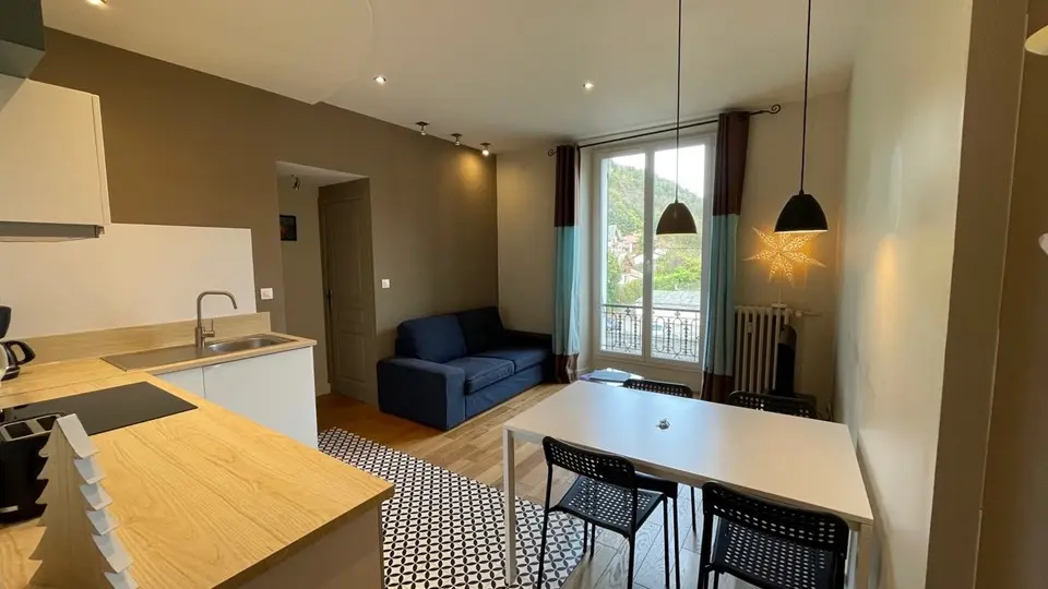 Cet appartement présente un espace de vie ouvert et  lumineux. La cuisine, équipée d'un plan de travail clair et de différents  électroménagers, est intégrée à l'espace principal. Sur la droite, une table à  manger et à gauche, un canapé bleu. 