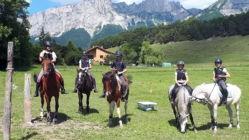 Un groupe d'adolescents à cheval se promènent dans une nature verdoyante, une montagne domine l'horizon contre le ciel bleu