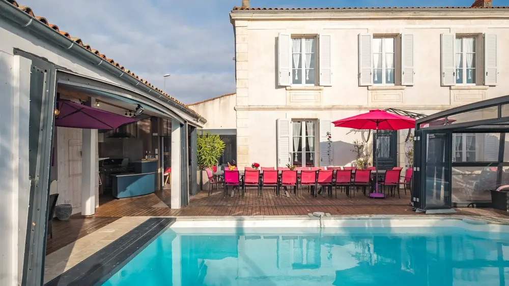 La piscine extérieure découverte, dans le fond table et chaises pour une quinzaine de personnes, la maison, à gauche une veranda ouverte.