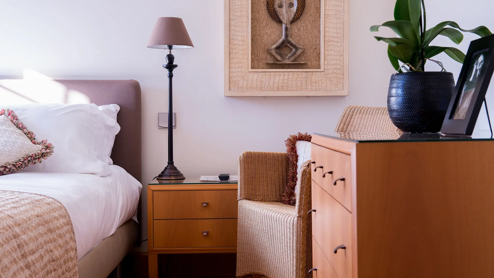 Cette chambre présente un lit double agrémenté de coussins et d'une couverture à motifs, une table de chevet en bois avec une lampe, et une commode assortie, le tout dans des tons clairs, avec une touche de verdure grâce à une orchidée.