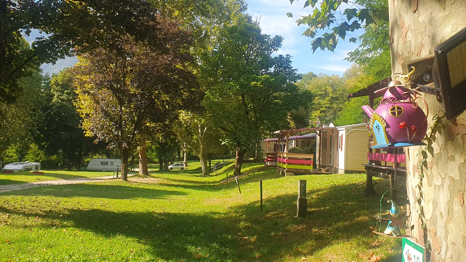 Un espace de camping verdoyant comprenant des parcelles avec des mobil-home séparées par des arbres. Le sol est principalement herbeux et des chemins semblent délimiter les différents emplacements.